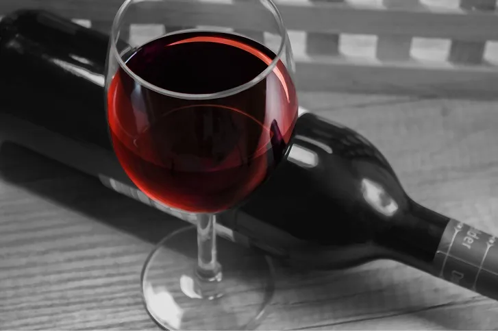 Copa de vino tinto en primer plano sobre una mesa de madera, con una botella de vino tumbada al fondo en blanco y negro.