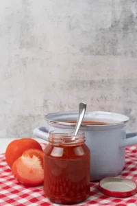 Tarro de cristal con salsa de tomate casera, acompañado de una olla azul, tomates frescos y una tapa metálica, sobre mantel de cuadros rojos.