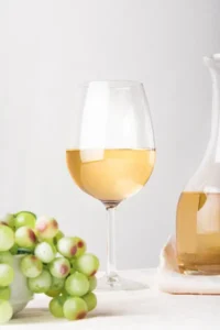 Copa de vino blanco junto a racimo de uvas verdes y botella de vidrio transparente sobre fondo claro.