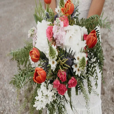 Ramo de novia con flores mixtas: tulipanes naranjas, rosas blancas, flores silvestres y follaje verde, sostenido por una mujer con vestido de encaje.