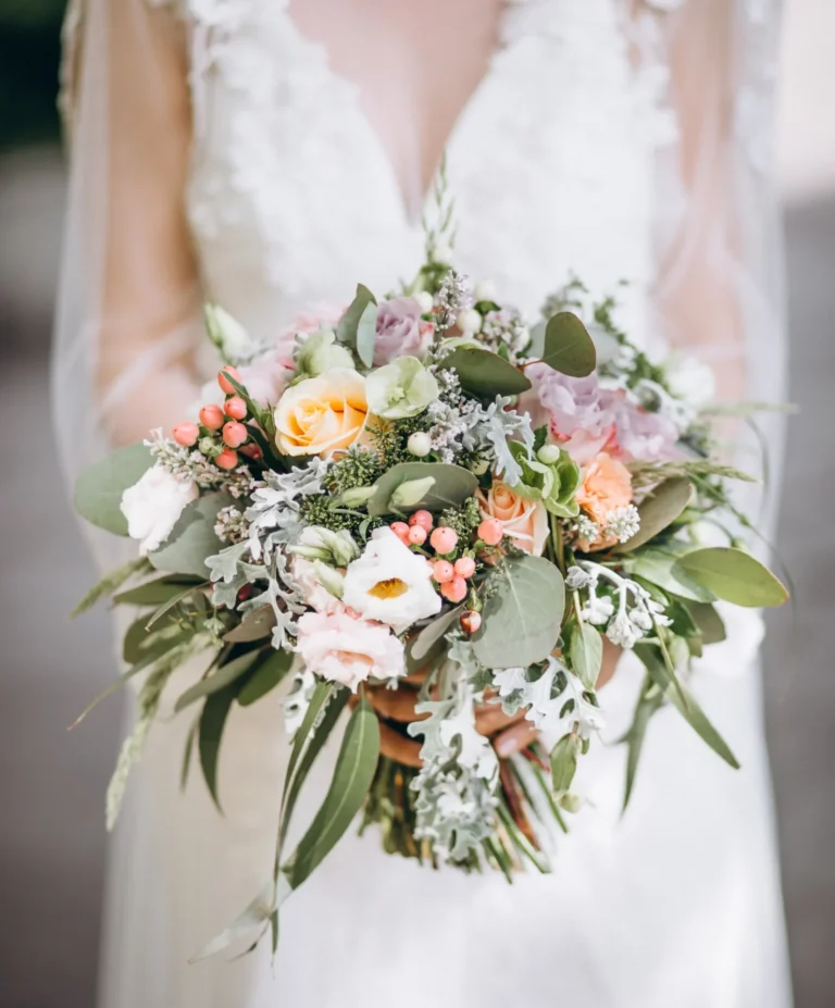 Novia sosteniendo un ramo de boda con flores en tonos pastel, follaje verde y bayas rosadas, en estilo silvestre y natural.