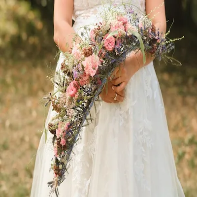Novia con vestido blanco sosteniendo un ramo de flores silvestres y rosas en forma alargada, en un entorno natural.