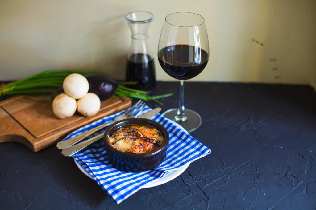 Plato horneado con queso fundido servido en cazuela de cerámica junto a copa de vino tinto, sobre mantel azul a cuadros, con berenjenas, cebolletas y champiñones de fondo.