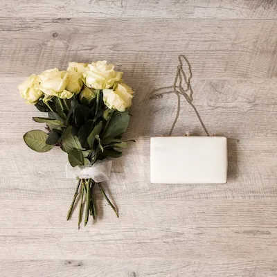 Ramo de rosas blancas atado con cinta de tul junto a un bolso de mano blanco con cadena, sobre fondo de madera clara.