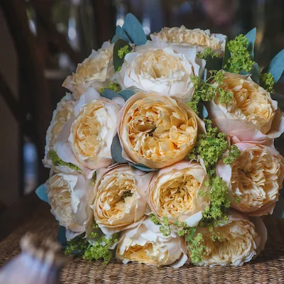Ramo compacto de flores tipo peonía en tonos crema y amarillo suave, con hojas verdes y detalles en eucalipto, sobre una mesa de bambú.