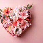 Caja en forma de corazón rellena de flores rosadas, blancas y naranjas sobre fondo rosa pastel, con un diseño romántico y delicado.