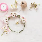 Imagen decorativa de una corona floral hecha a mano con margaritas blancas, cinta rosa y cuerda de yute sobre fondo claro de madera.